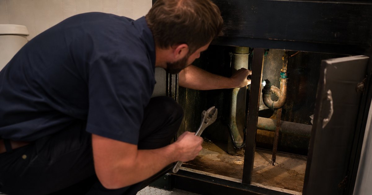 A plumber crouched under an open bathroom vanity in an older rental unit, flashlight beam on damp copper pipes and a water stained cabinet floor, adjustable wrench in one hand, dim indoor light and a towel on the tile